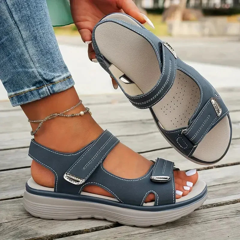 Blue sandals with velcro straps worn by a person on a wooden floor.